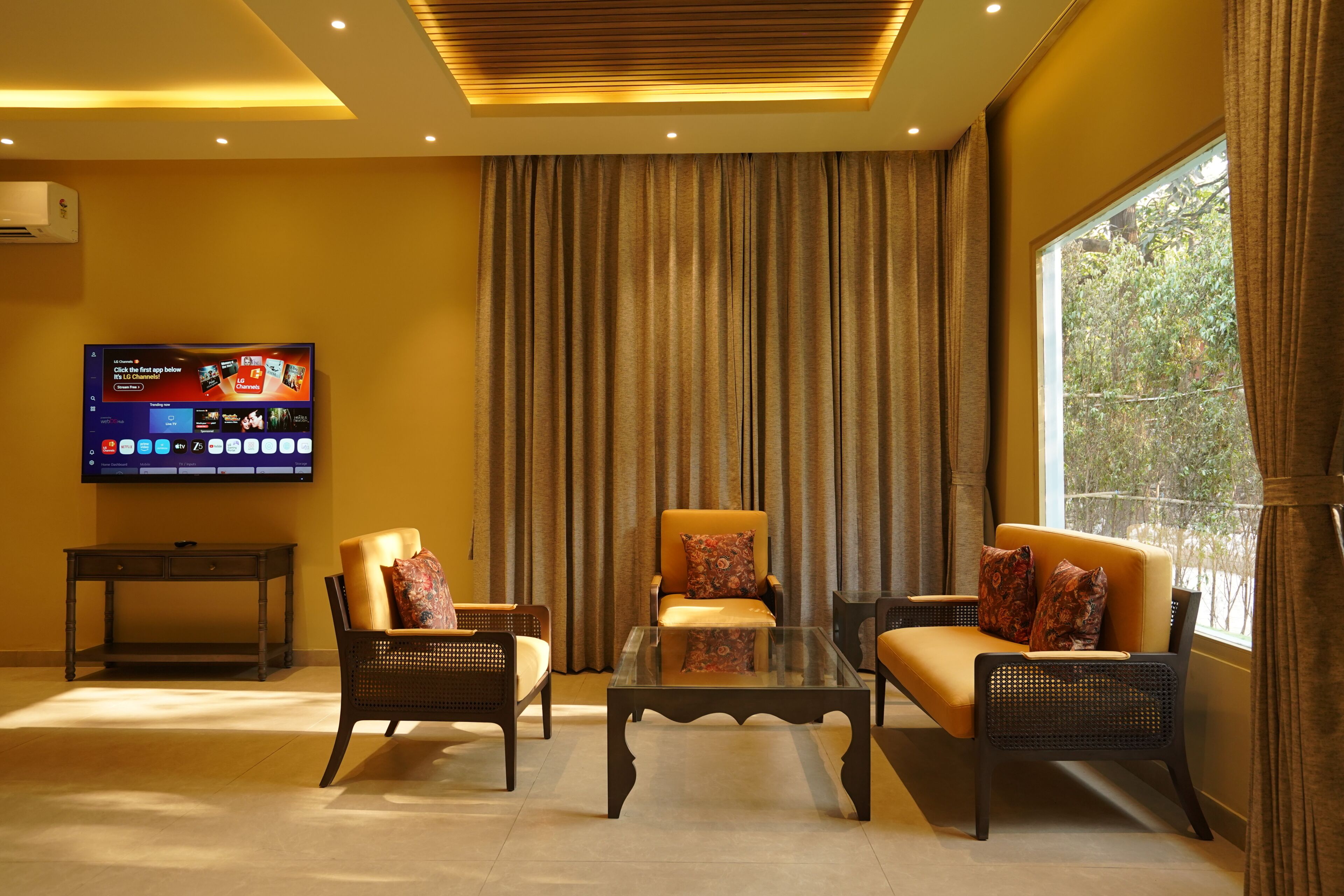 A living room with a flat-screen TV, two armchairs, a coffee table, and a large window with curtains.