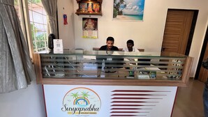En receptionsdisk med en glasvägg, två personer sittande bakom den, en trähylla med religiösa föremål, en kalender och en inramad tavla med ett strandmotiv.