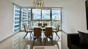 A modern dining area with a large window offering a city view, a contemporary chandelier, and a clean, minimalist design. - Gold Tower - Bright 3 BDR Apt in Costa del Este (Panama City)