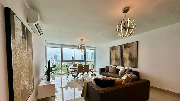 A modern living room with a large window, a sofa, a dining area, and artwork on the walls. - Gold Tower - Bright 3 BDR Apt in Costa del Este (Panama City)