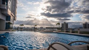 A rooftop swimming pool with lounge chairs and palm trees, surrounded by high-rise buildings and a cloudy sky. - Gold Tower - Bright 3 BDR Apt in Costa del Este (Panama City)