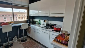 A compact kitchen with white cabinets, a window with a view of buildings, a small bar stool, and a red pot on the counter. - "BlueSky" air-conditioning-wi-fi garage- 5m from town center.  (Aix-en-Provence)