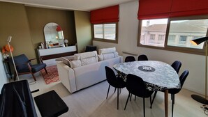 A modern living room with a sofa, a dining table with chairs, a small side table, a mirror, and a window with red curtains. - "BlueSky" air-conditioning-wi-fi garage- 5m from town center.  (Aix-en-Provence)