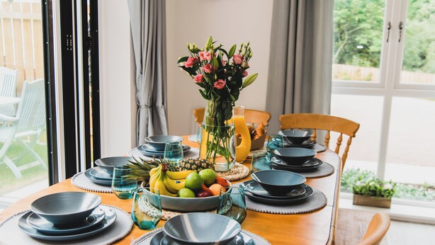 A dining table set with black plates, bowls, and a fruit bowl. There is a vase with flowers and a glass of orange juice.