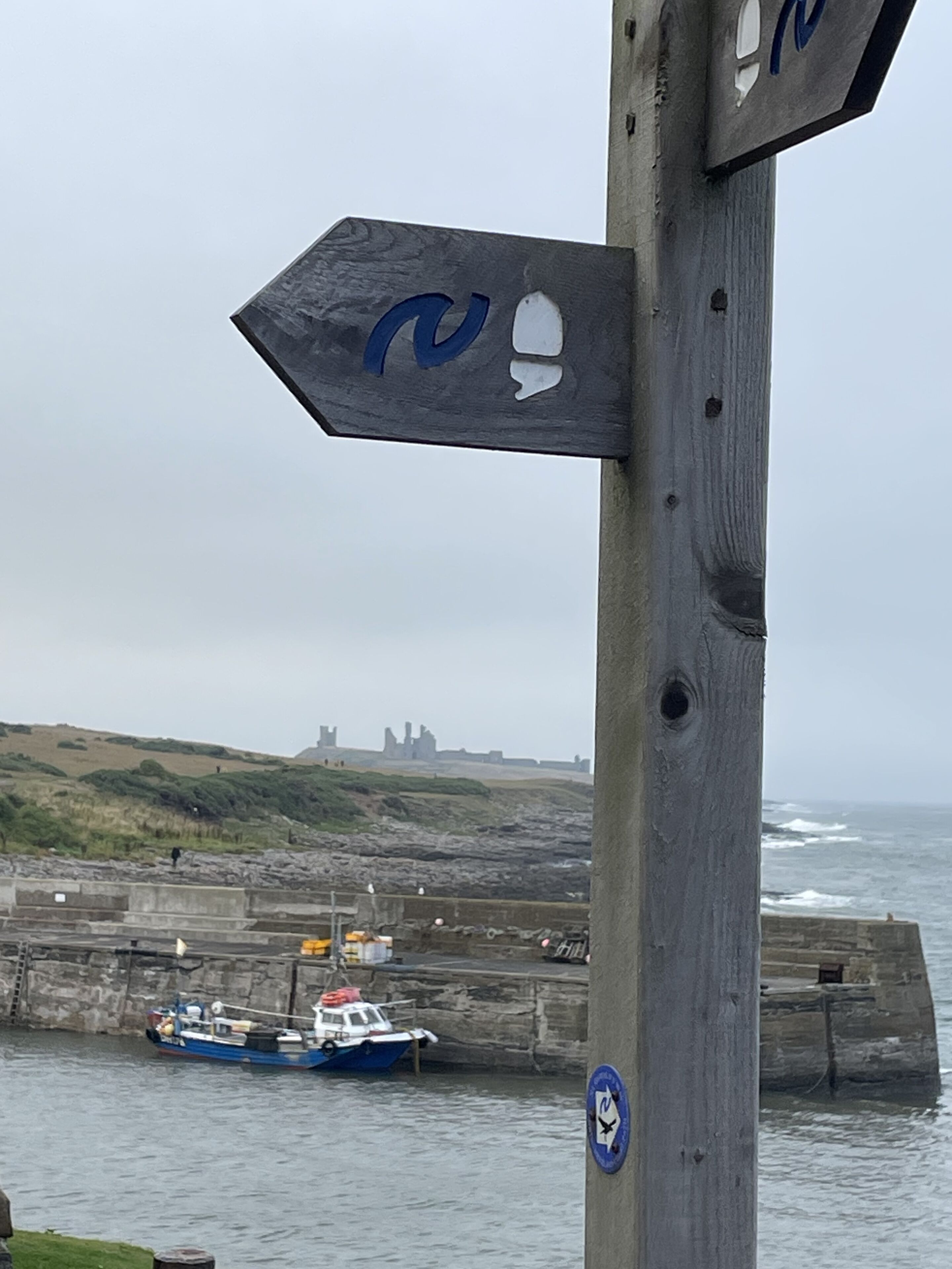 A wooden signpost with a blue arrow and a white arrow pointing in different directions, overlooking a coastal area with a boat docked near a stone wall and a castle in the background.