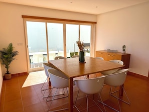Un coin repas avec une table en bois, des chaises blanches, une plante en pot et une vue dégagée grâce à de grandes portes vitrées.