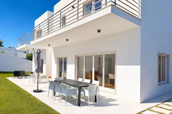 A modern two-story house with a patio, outdoor seating, and a large sliding glass door.