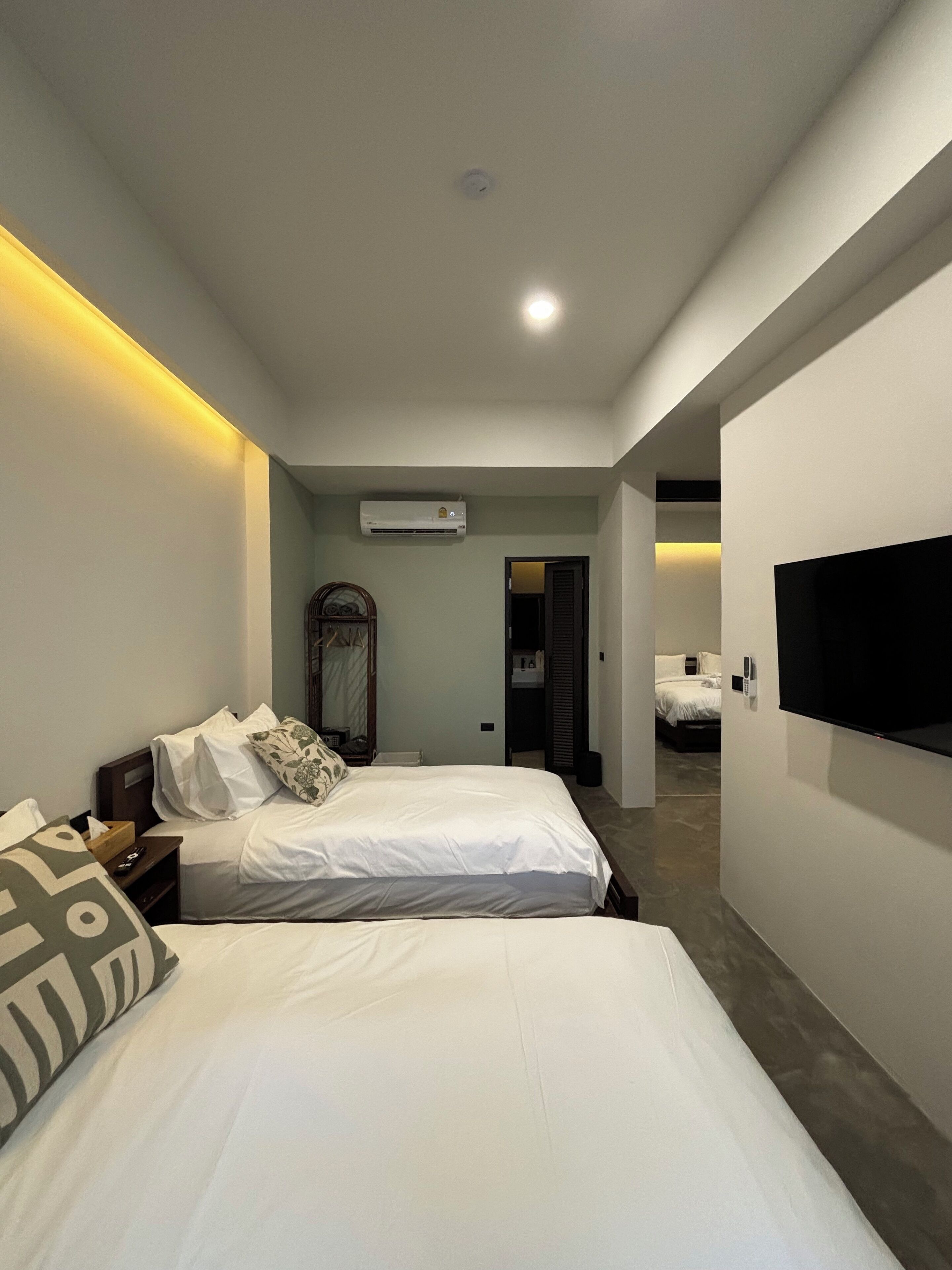 A modern hotel room with two beds, a flat-screen TV, an air conditioning unit, and a hallway leading to another room.