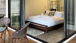 Een moderne slaapkamer met een glazen deur die leidt naar een balkon, een bed met witte lakens en een zithoek met rieten stoelen.
