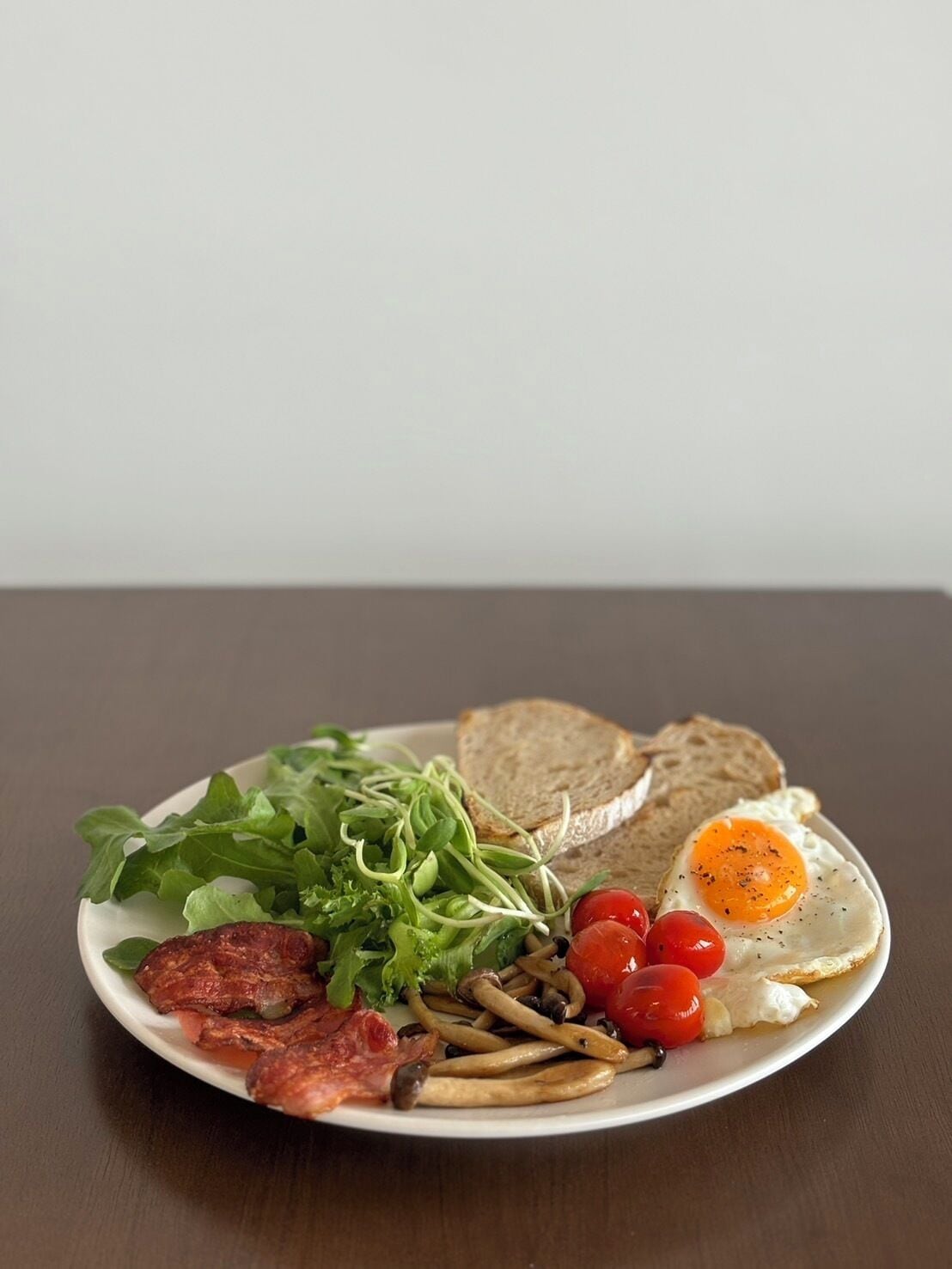 A plate with a fried egg, bacon, mushrooms, cherry tomatoes, lettuce, and slices of bread.