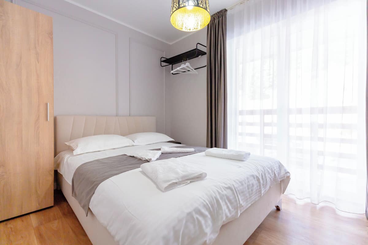 A neatly made bed with white linens, a wooden wardrobe, a hanging clothes rack, and a window with sheer curtains.