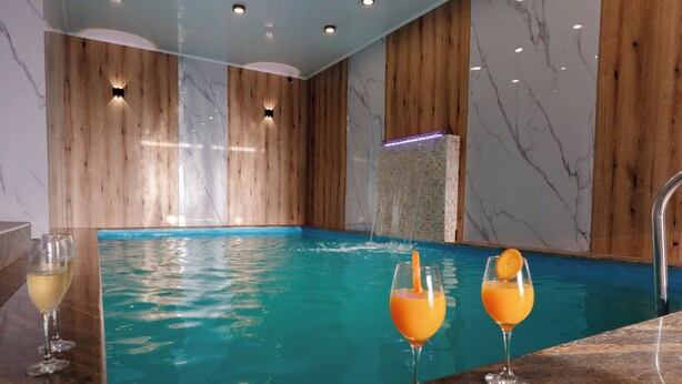 An indoor swimming pool with wooden walls and marble accents, two glasses of orange juice, and a glass of white wine on a counter. - Complex Veverita  (Novaci)