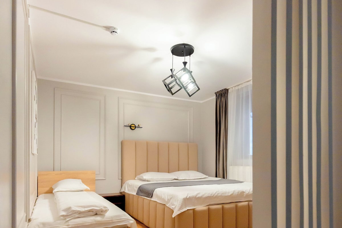 A bedroom with two beds, wooden flooring, a ceiling light fixture, and a window with curtains.