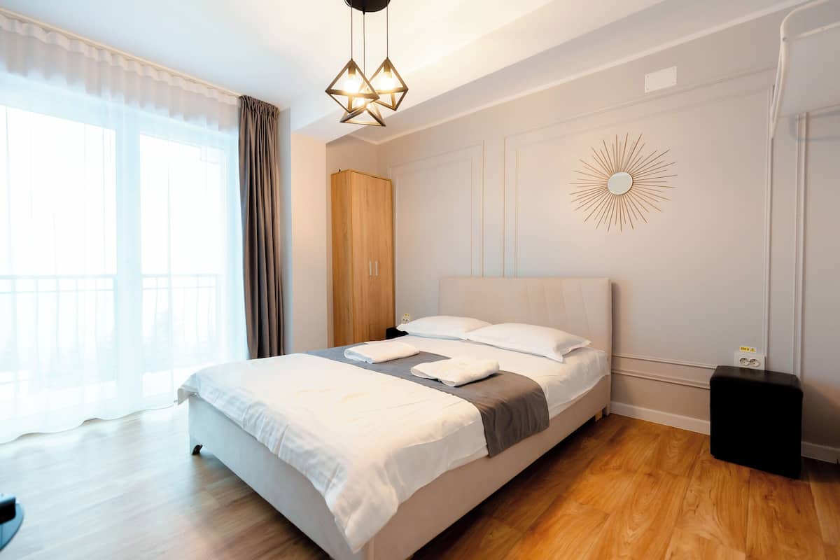 A neatly made bed with white linens, a wooden wardrobe, a sunburst wall decoration, and a modern pendant light.