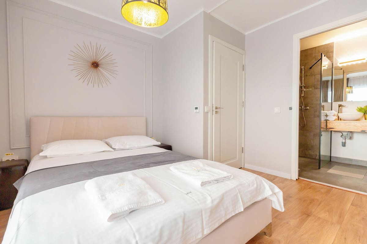 A modern hotel room with a large bed, a bedside table, a wall-mounted light fixture, and a bathroom visible through an open door.