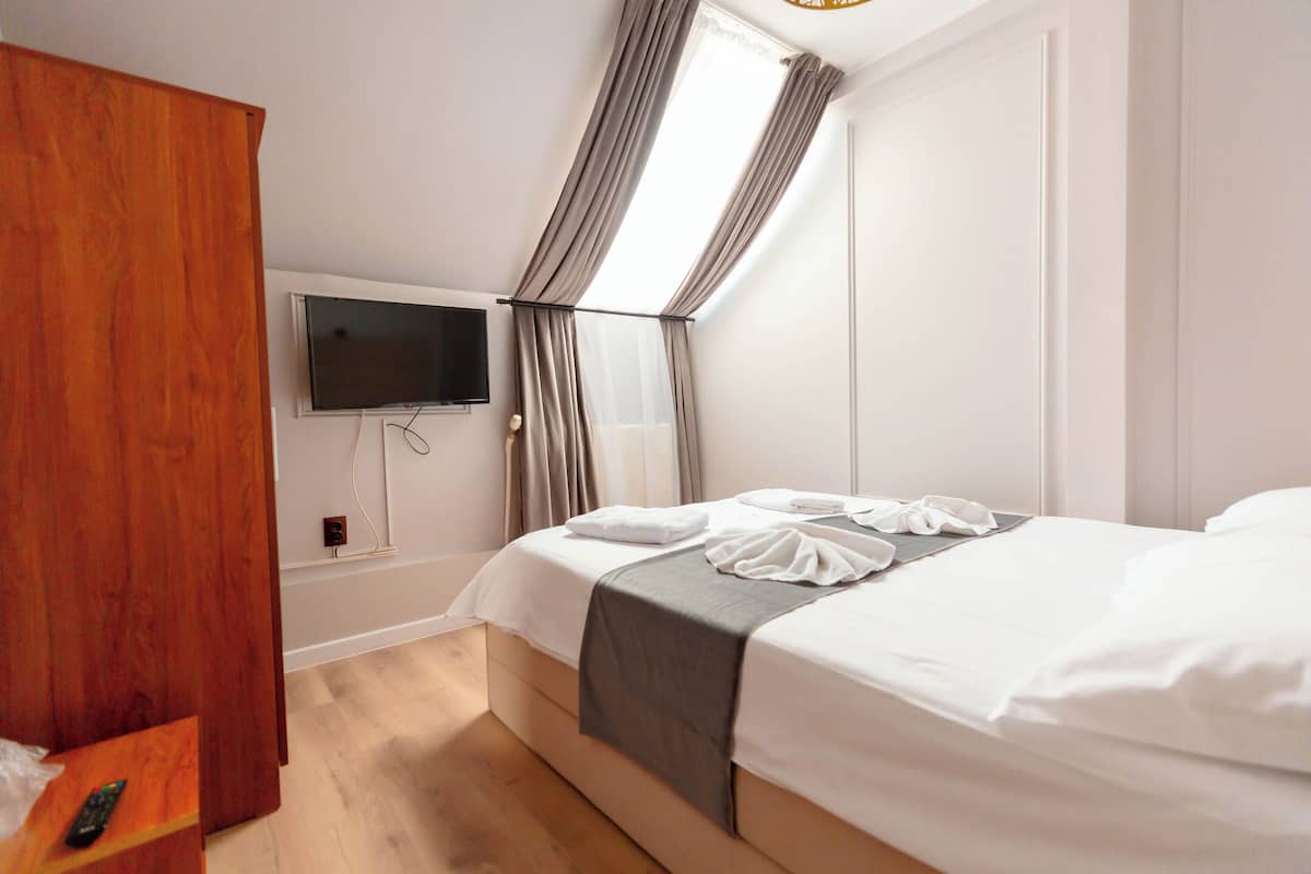 A hotel room with a bed, a TV mounted on the wall, a wooden wardrobe, and a window with curtains.