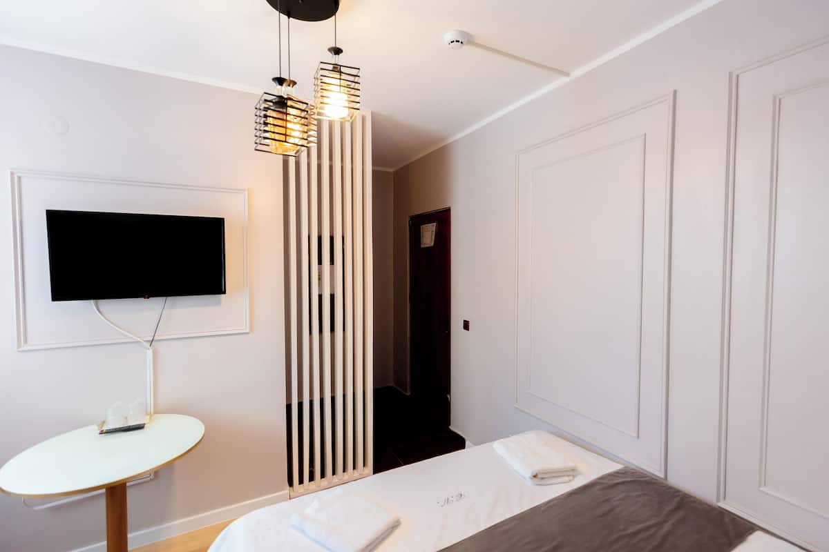 A modern hotel room with a flat-screen TV mounted on the wall, a bedside table with a remote control, a bed with a grey blanket, and a decorative light fixture above the bed.