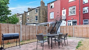 Eine Terrasse mit Grill, Tisch und Stühlen, umgeben von einem Holzzaun und Gebäuden im Hintergrund.