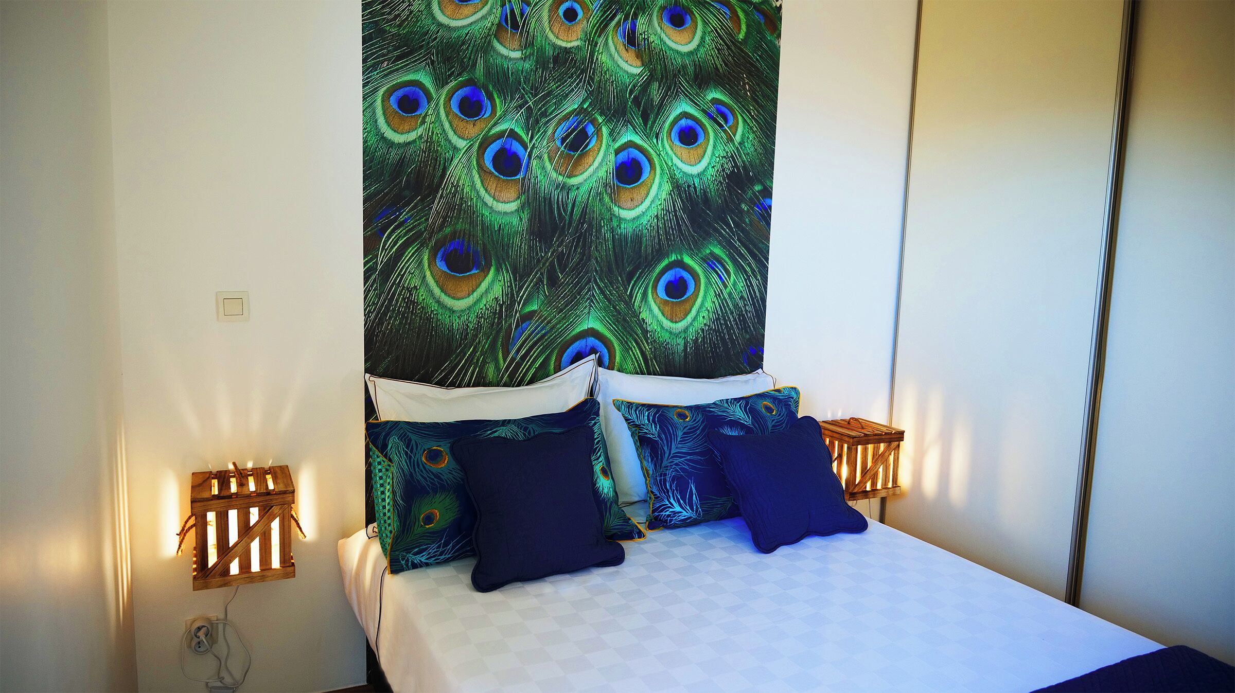 A bedroom with a bed, peacock-themed wall tapestry, and two bedside tables with lamps.