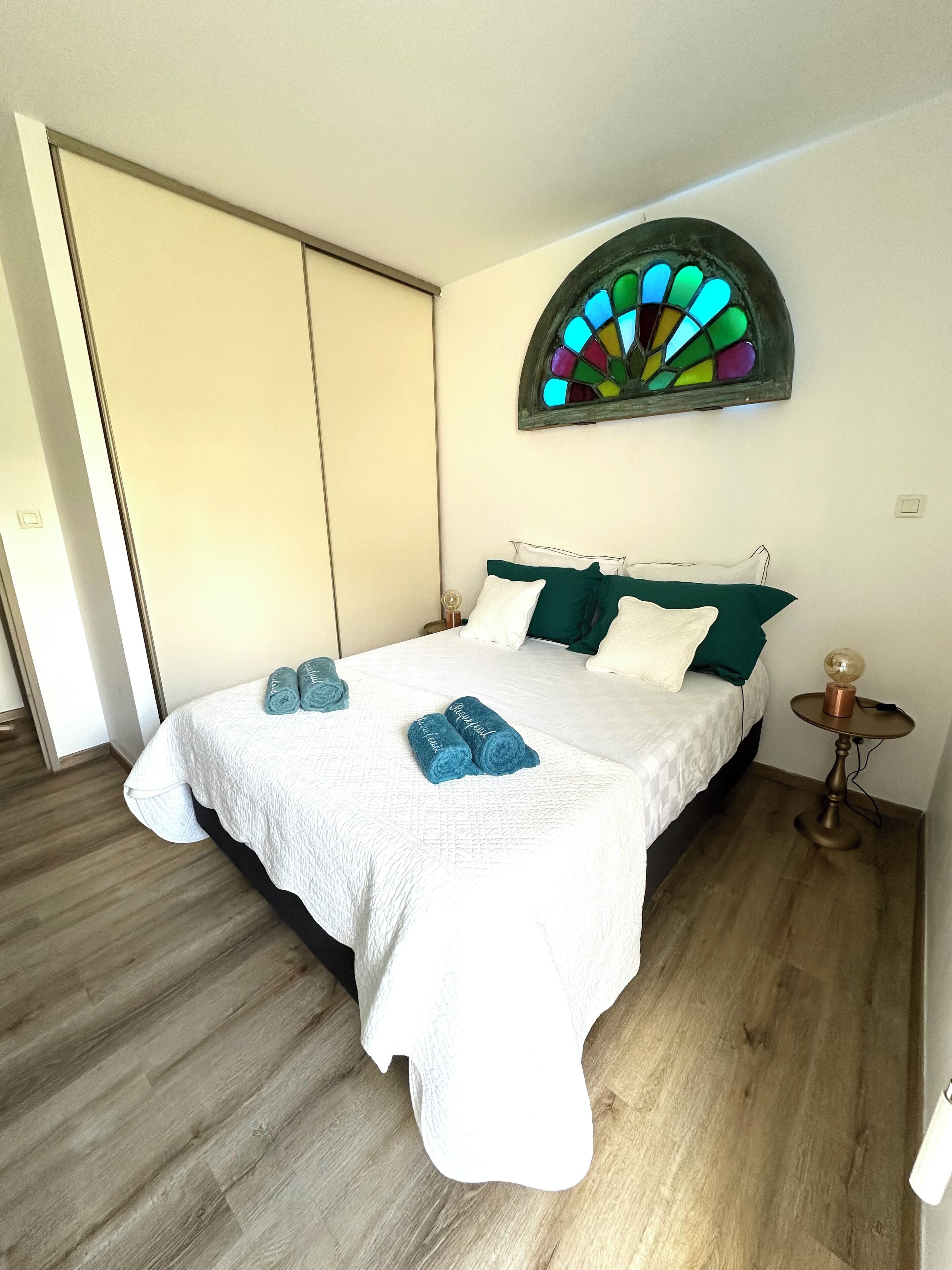 A bedroom with a bed, a nightstand, a wall-mounted lamp, and a stained glass window.
