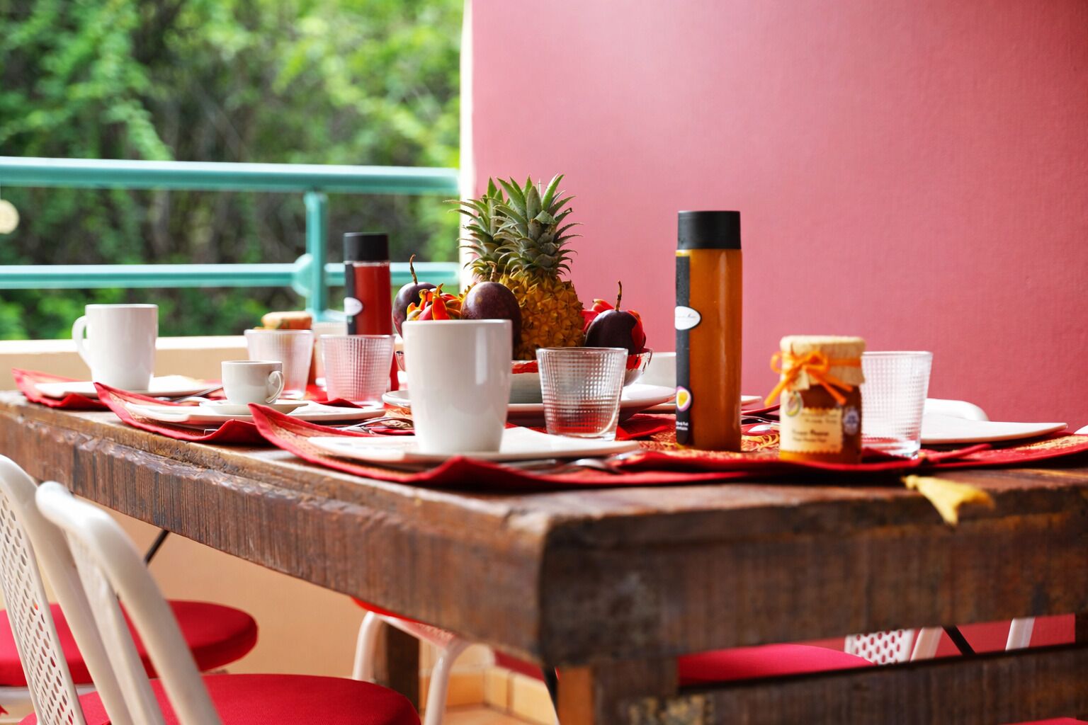 A table set with white cups, a pineapple, a bottle, and a jar, with a red tablecloth and a view of greenery outside.