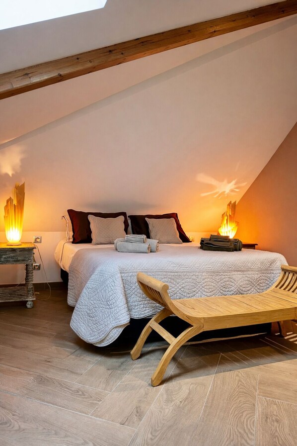 A bedroom with a sloped ceiling, a bed with a white quilt, two bedside lamps, a wooden bench, and a small table with a lamp.