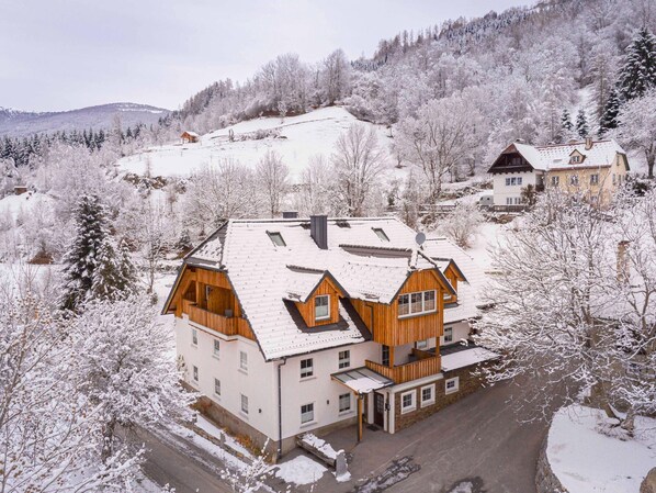 A multi-story wooden house with a sloped roof, surrounded by snow-covered trees and a hilly landscape.