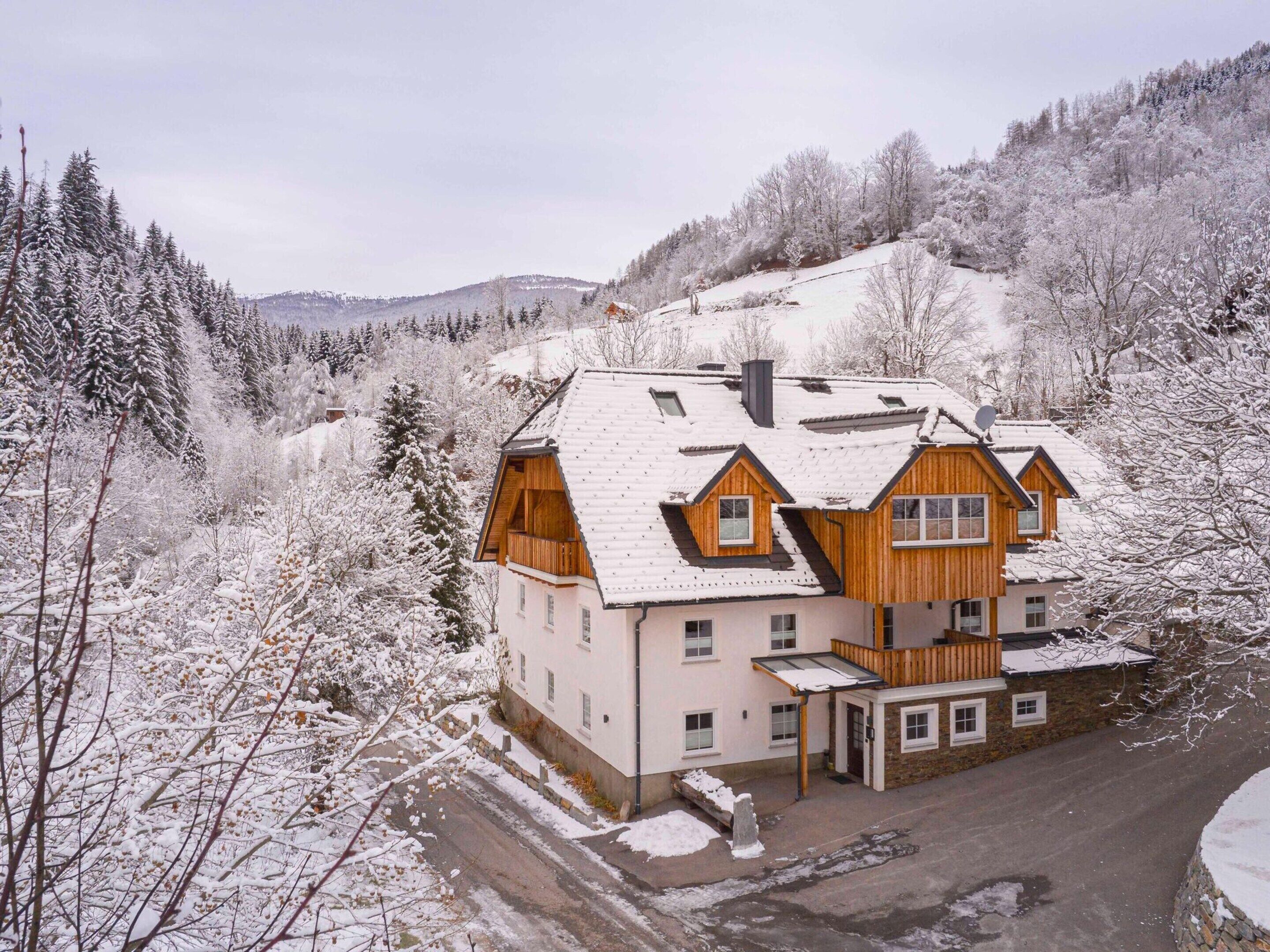 Um chalé de madeira de vários andares com telhado inclinado, rodeado por árvores e colinas cobertas de neve.