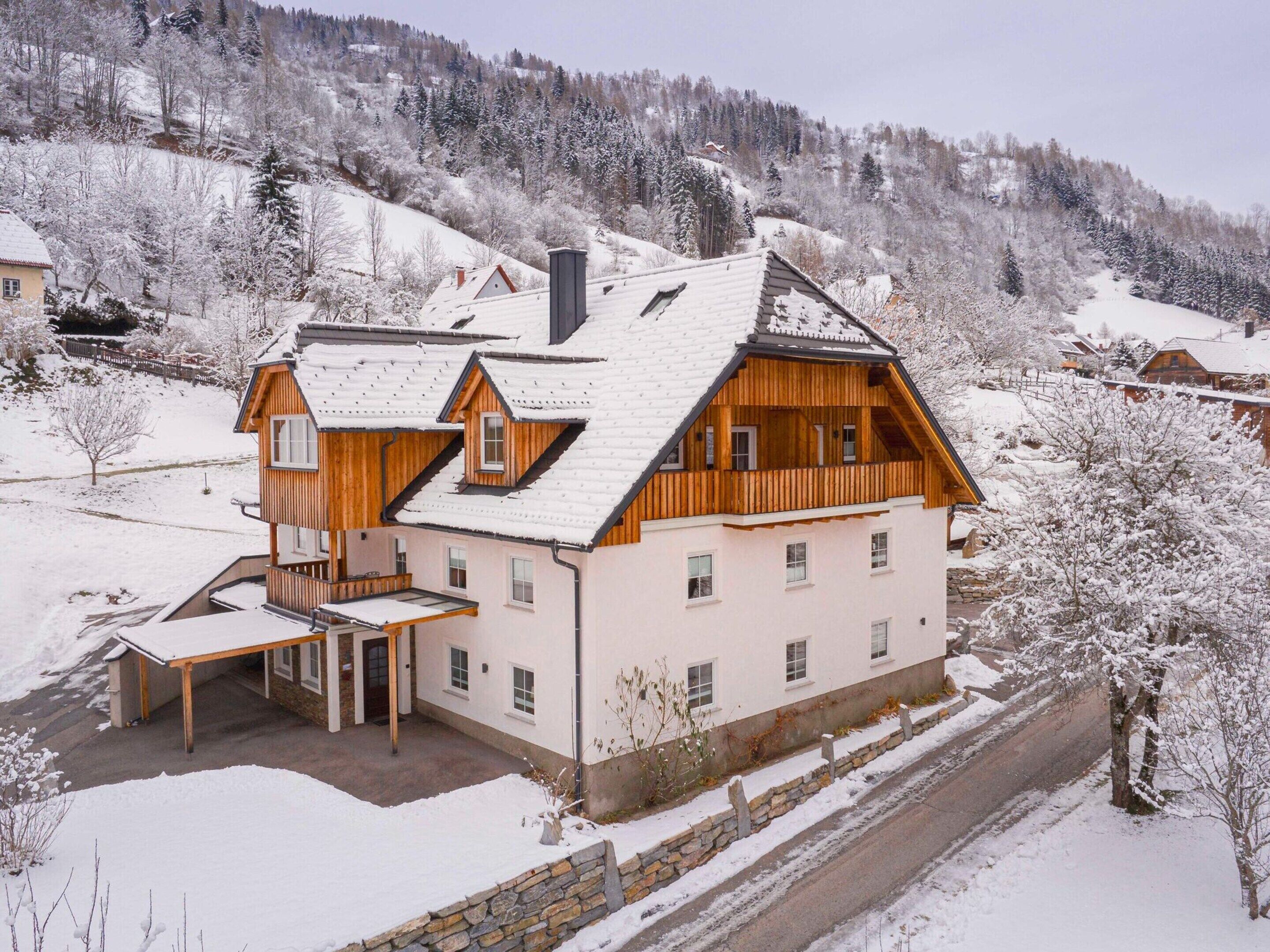 A two-story wooden house with a sloped roof, a balcony, and a stone wall surrounding the property.