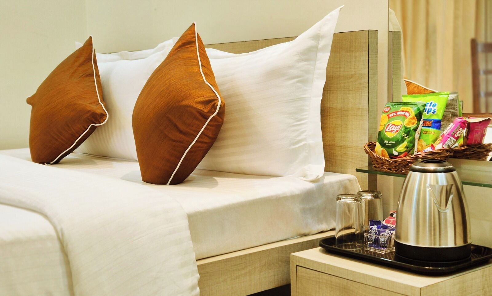 Quarto de hotel com cama, duas almofadas castanhas, lençóis brancos, um suporte com lanches, chaleira e um cesto de petiscos.
