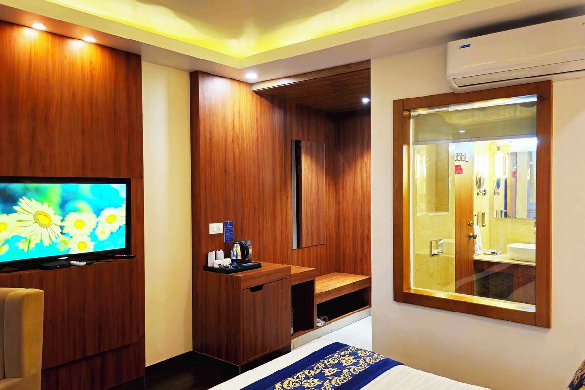A hotel room with a TV, wooden panel walls, a minibar, a bed with a blue and gold bedspread, and an air conditioning unit.