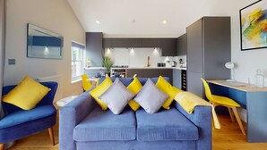 A modern living room with a blue sofa and yellow cushions, a dining area with a wooden table and yellow chairs, and a kitchen in the background.