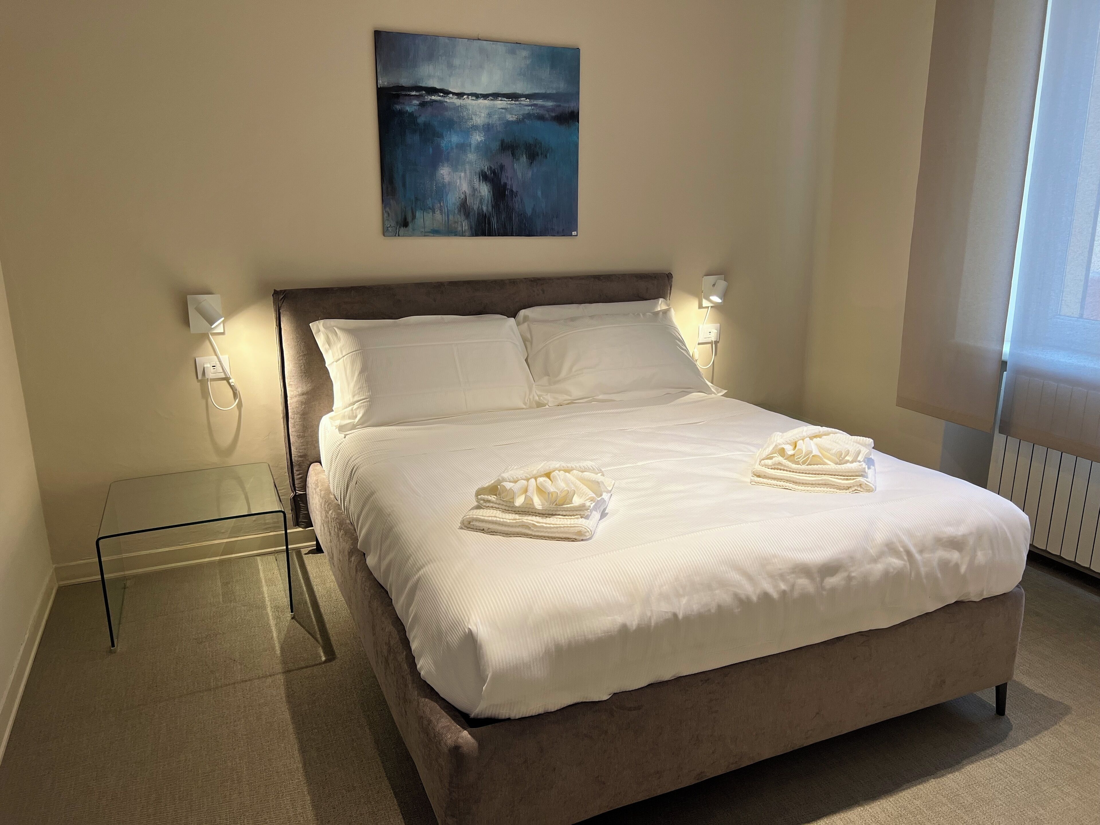 Una cama bien hecha con ropa y toallas blancas, un cuadro encima y una mesita de cristal al lado.