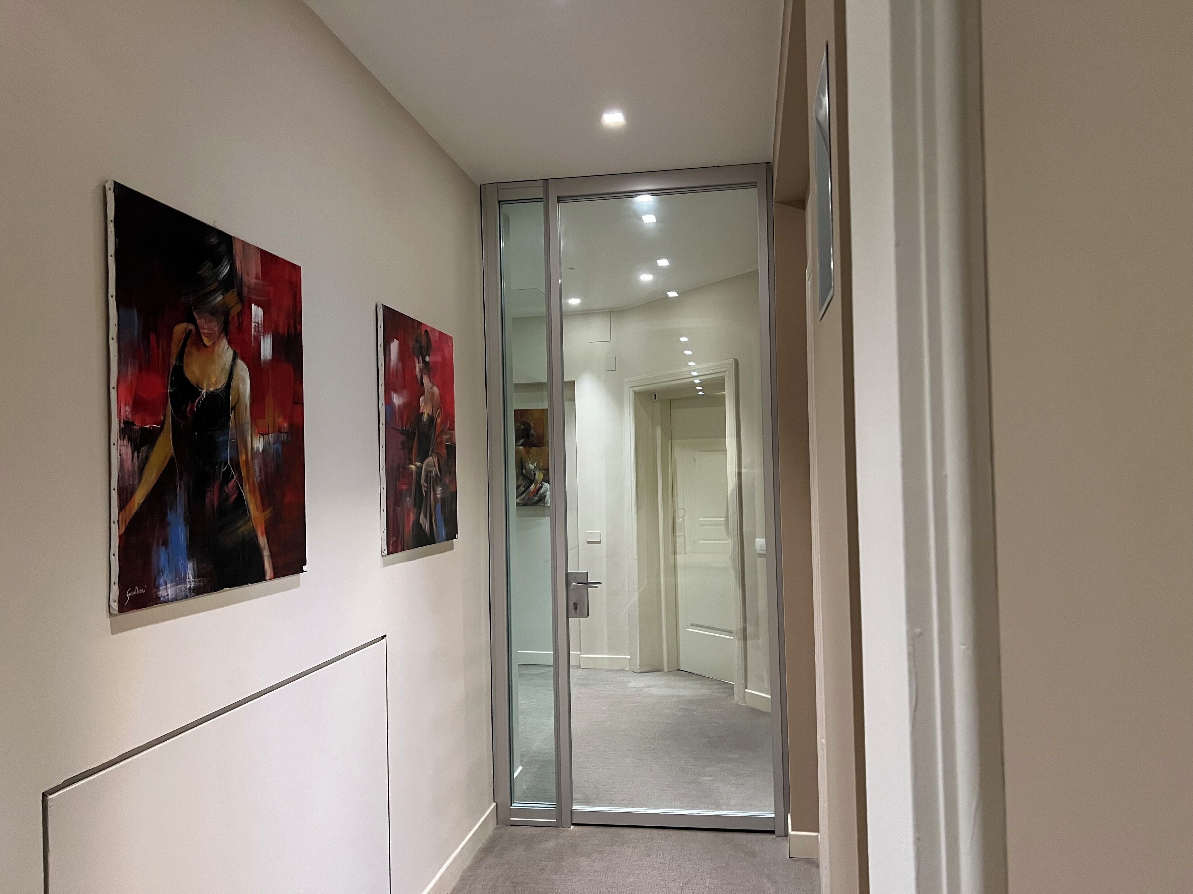 A hallway with two paintings on the wall, a glass door leading to another room, and a carpeted floor.