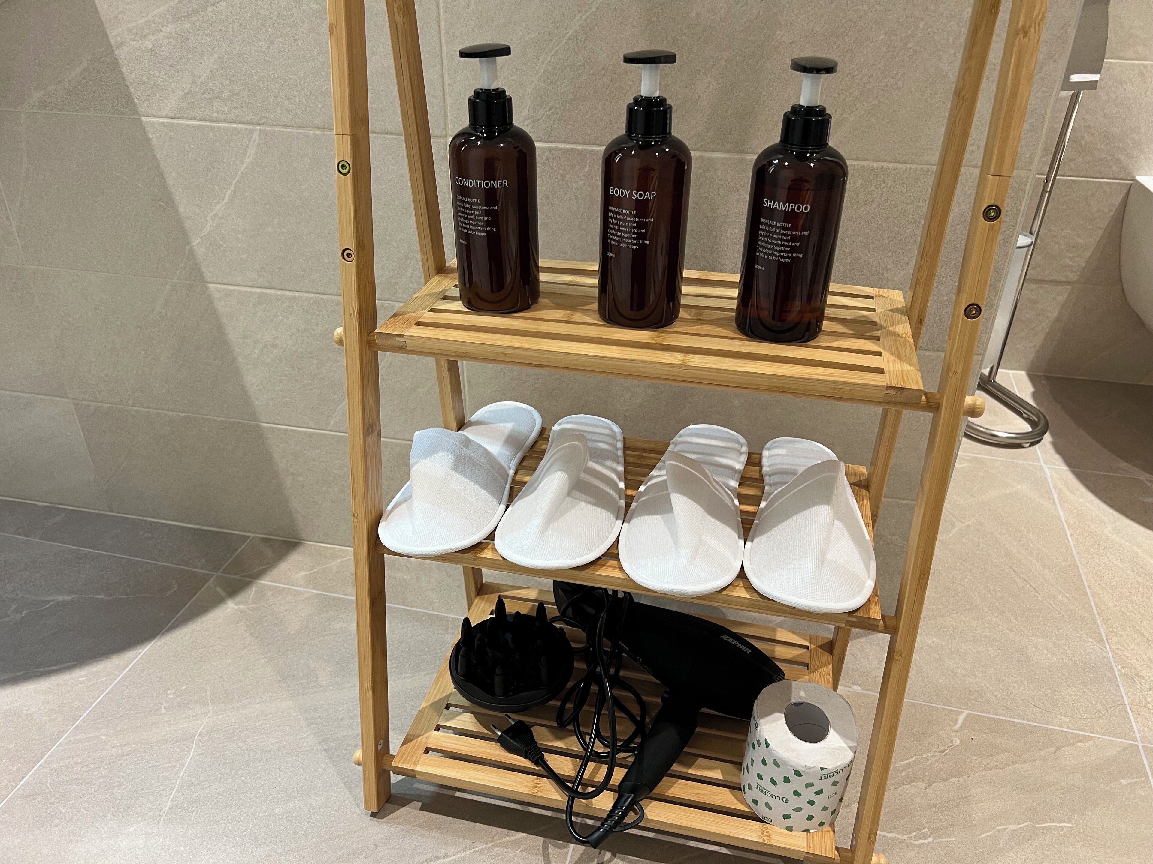 A wooden shelf with three bottles of body soap, a pair of white slippers, a black hairdryer, and a roll of toilet paper.