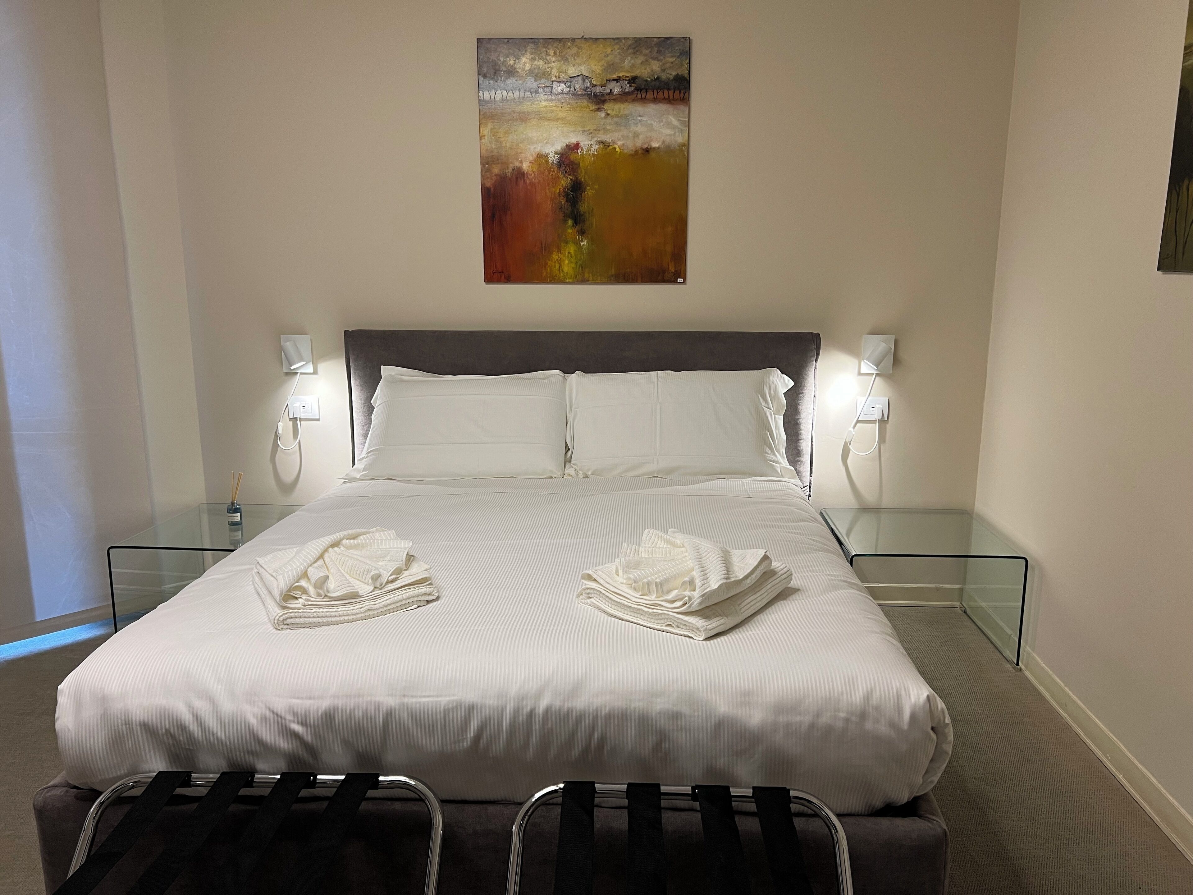 A neatly made bed with white linens, two bedside tables with lamps, and a painting above the bed.