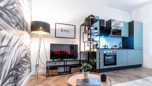 Een moderne woonkamer met een flatscreen televisie, een salontafel met een laptop, een vloerlamp en een keuken met kasten en apparatuur.
