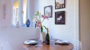 Een ronde eettafel met witte stoelen, een vaas met bloemen, een fles champagne en ingelijste foto's aan de muur.