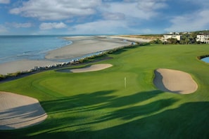 Campo de golf con búnkeres, una bandera y vista al océano y la playa.
