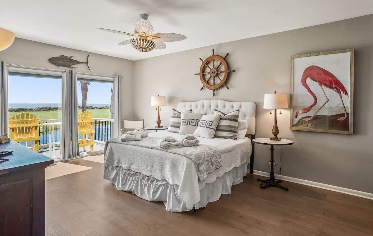 A bedroom with a bed, a ceiling fan, a ship's wheel, a framed painting, and a view of the outdoors.