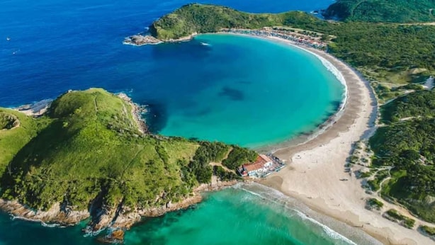 An aerial view of a coastal area with a curved beach, lush green hills, and clear blue waters. - Tropical Beach / Center - Cabo Frio (Cabo Frio)