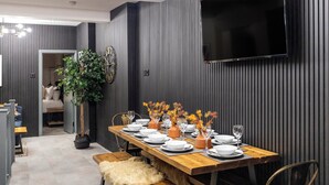 A dining area with a long wooden table set for six, flanked by dark vertical paneling and a mounted TV.