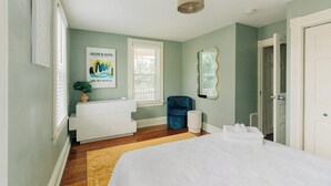A bedroom with a bed, a white dresser, a blue chair, a mirror, and a window with blinds. - The Garden House (Lancaster)
