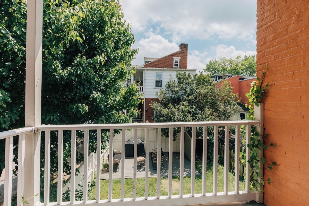 Ein Balkon mit weißer Reling blickt auf einen Hinterhof mit Rasenfläche, einer Terrasse mit Gartenmöbeln und einer Ziegelmauer.