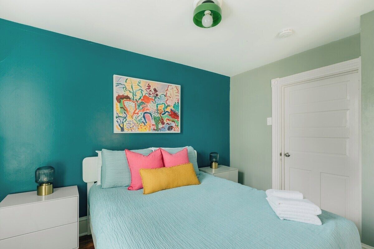 Une chambre avec un mur de couleur turquoise, un lit agrémenté d’oreillers roses et jaunes, un tableau au-dessus du lit et une porte blanche.