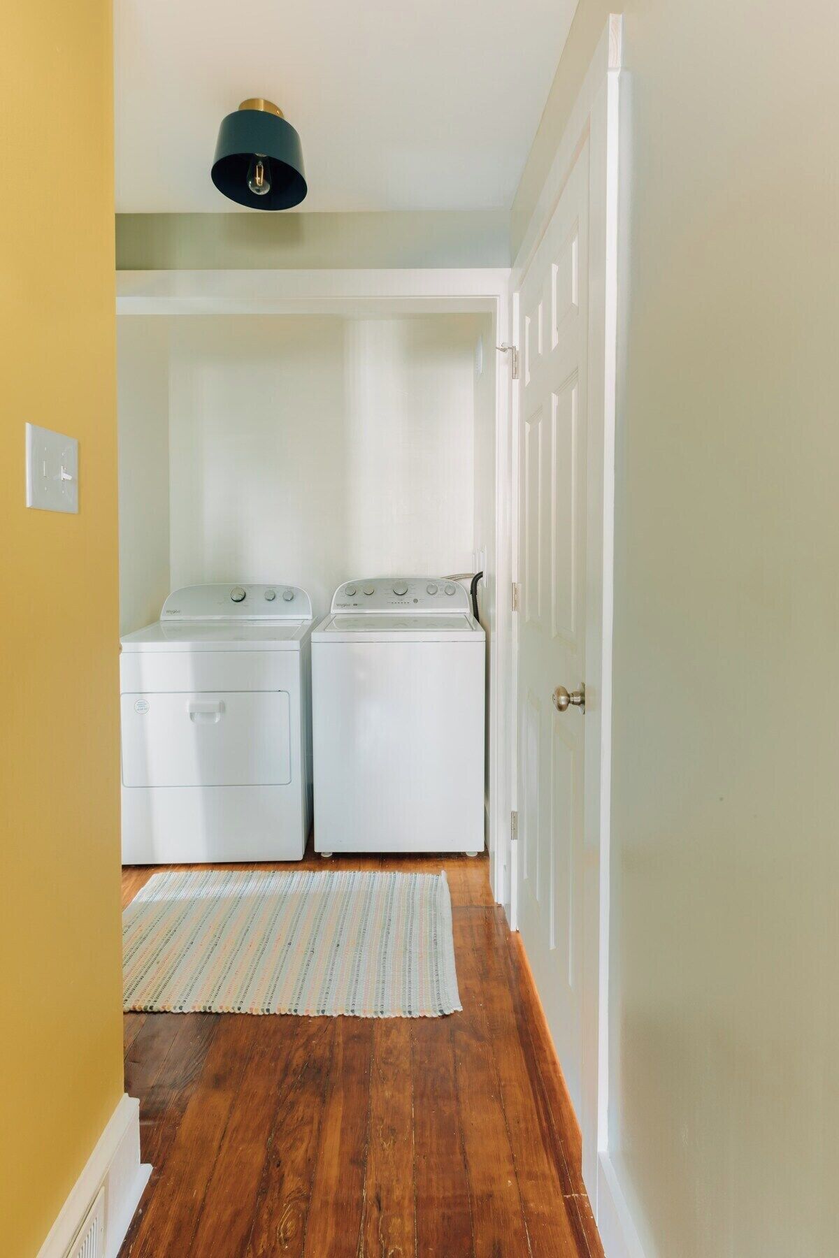 A laundry room with two white front-loading washers and dryers, a striped rug on the wooden floor, and a door leading to another room.