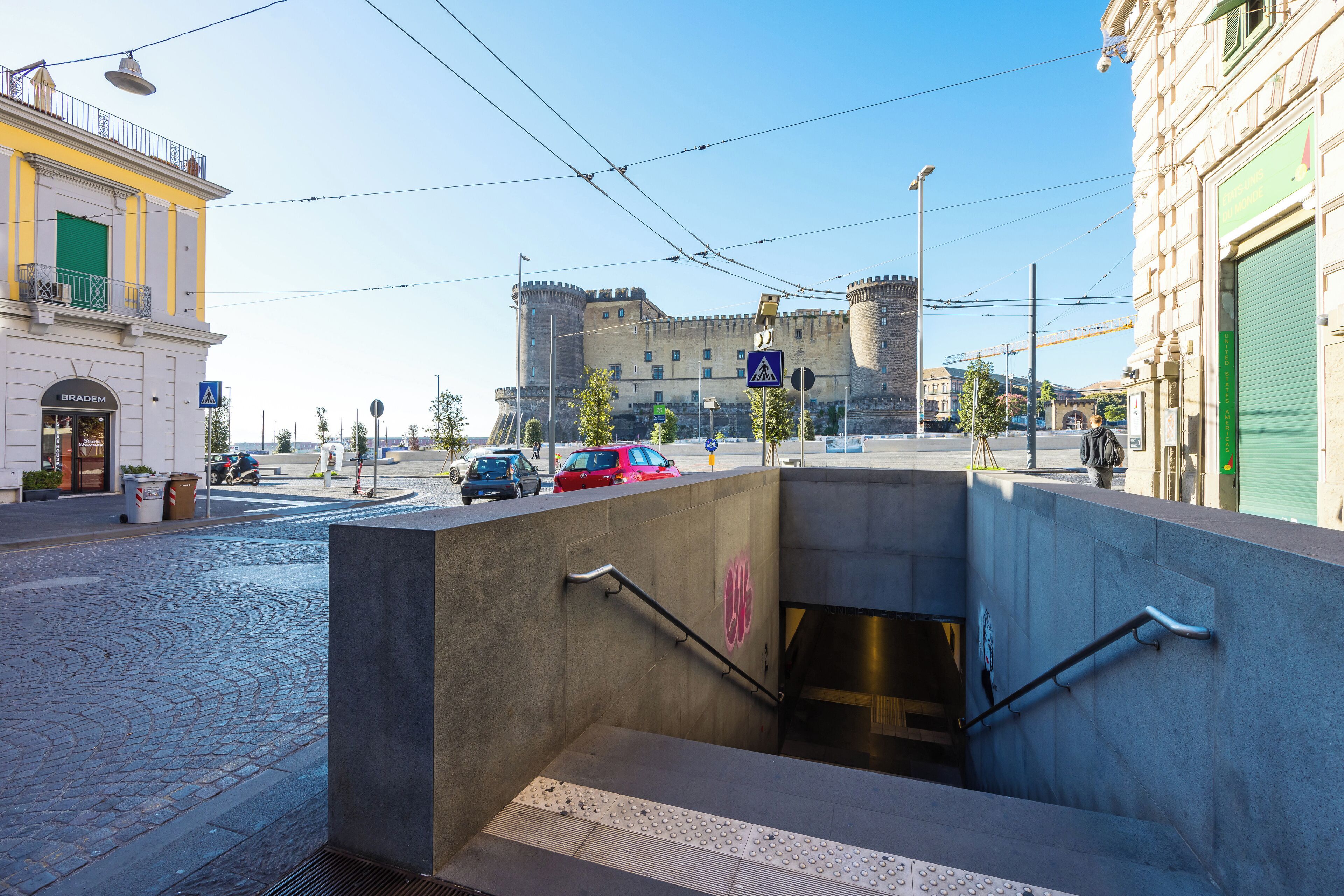 A street view with a castle in the background, a tram line overhead, and a building with a green shutter.