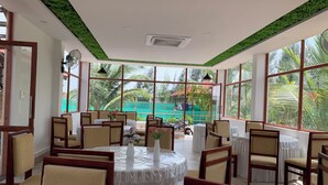 Un restaurant avec des tables rondes recouvertes de nappes blanches et des chaises en bois. De grandes fenêtres offrent une vue sur des palmiers et une clôture verte à l’extérieur.