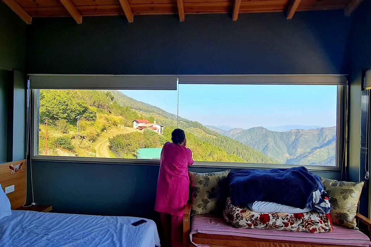 Una habitación con una cama, un ventanal con vistas a la montaña y una persona de pie junto a la ventana.
