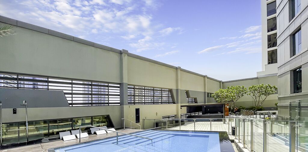 A rooftop pool area with a modern building in the background, glass railings, and a clear sky.
